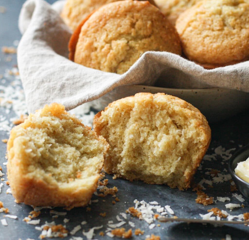 Coconut Corn Muffins in a basket, with a sliced muffin in the foreground