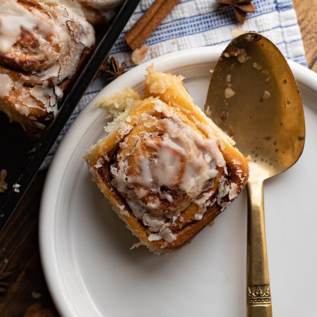 One classic cinnamon roll on a white plate with a gold serving spoon