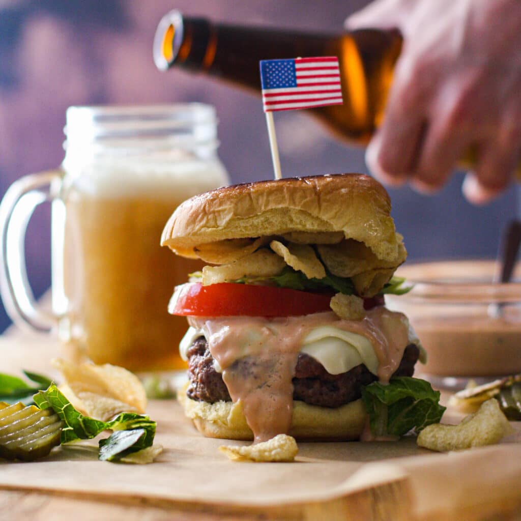 Cheeseburger with lettuce, tomato, potato chips and special sauce with someone pouring a beer in the background.