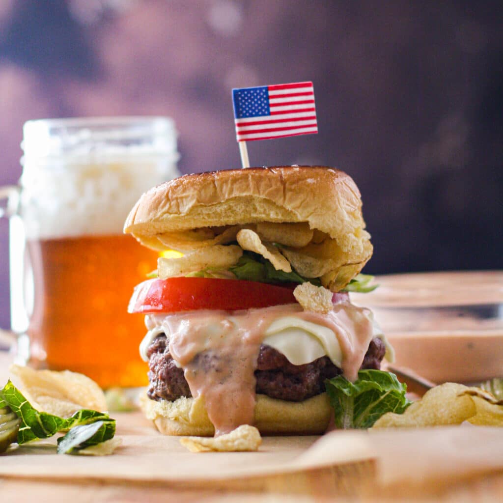 Cheeseburger with lettuce, tomato and potato chips with an American flag toothpick sticking up from the top