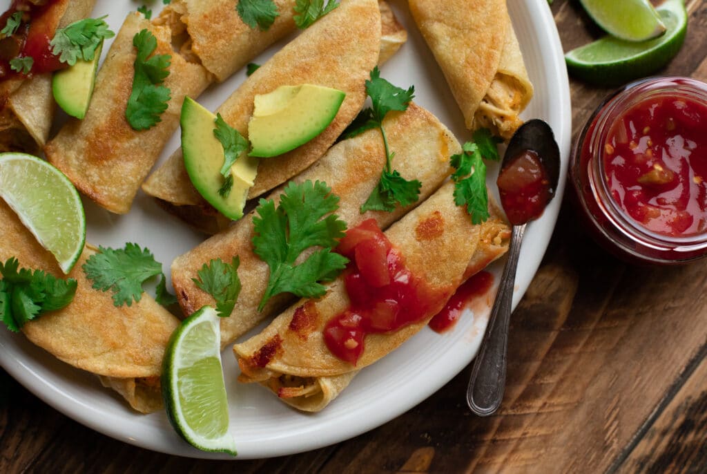 Baked Chicken Taquitos on a white plate, garnished with lime, cilantro and avocado