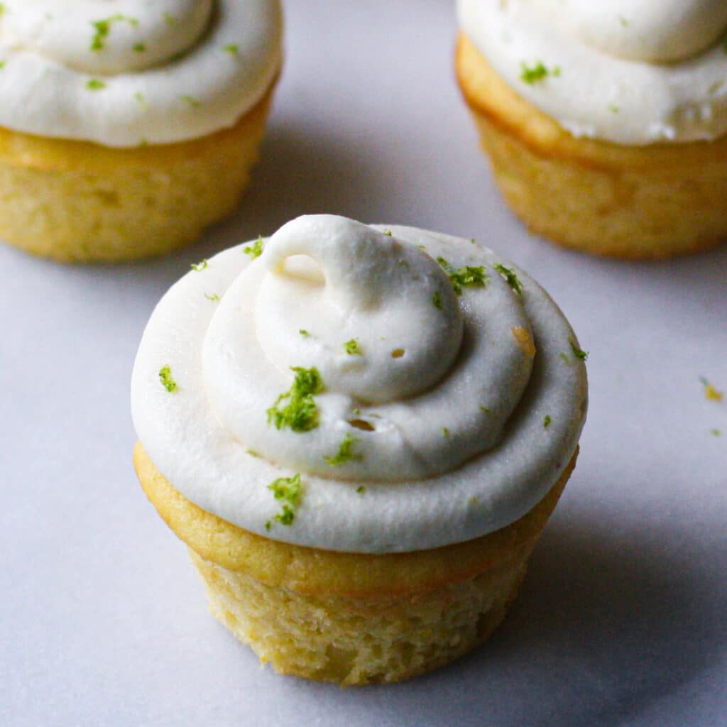 Yellow cupcake with white frosting swirled on top and sprinkled with green lime zest against a pale gray surface