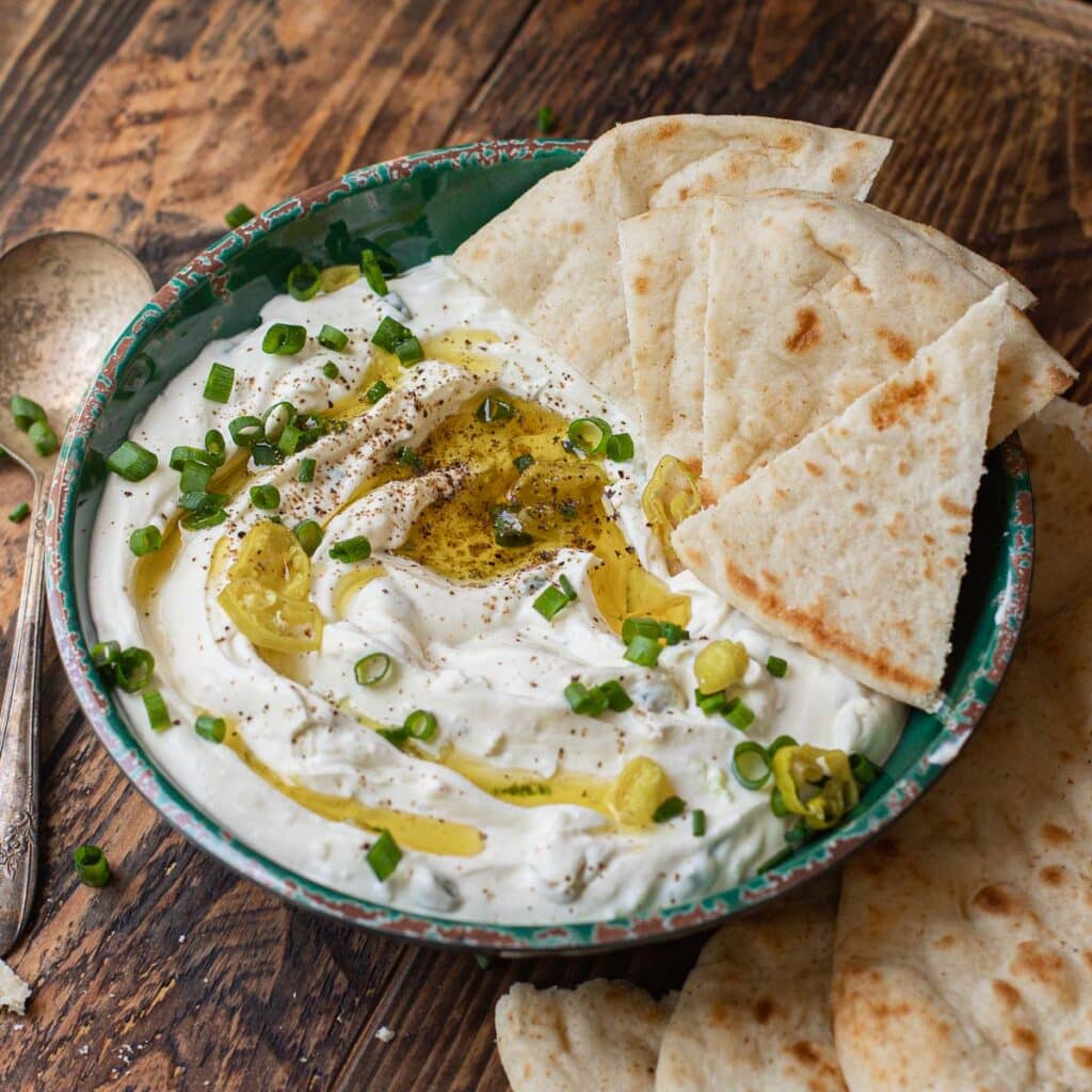 A bowl of Greek feta dip, drizzled with olive oil and garnished with scallions, pepperoncini peppers and pita bread