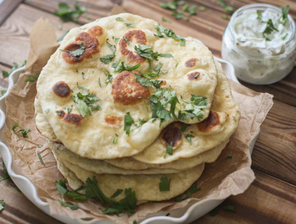 a pile of naan on a white plate on a wooden table with raita in the background.