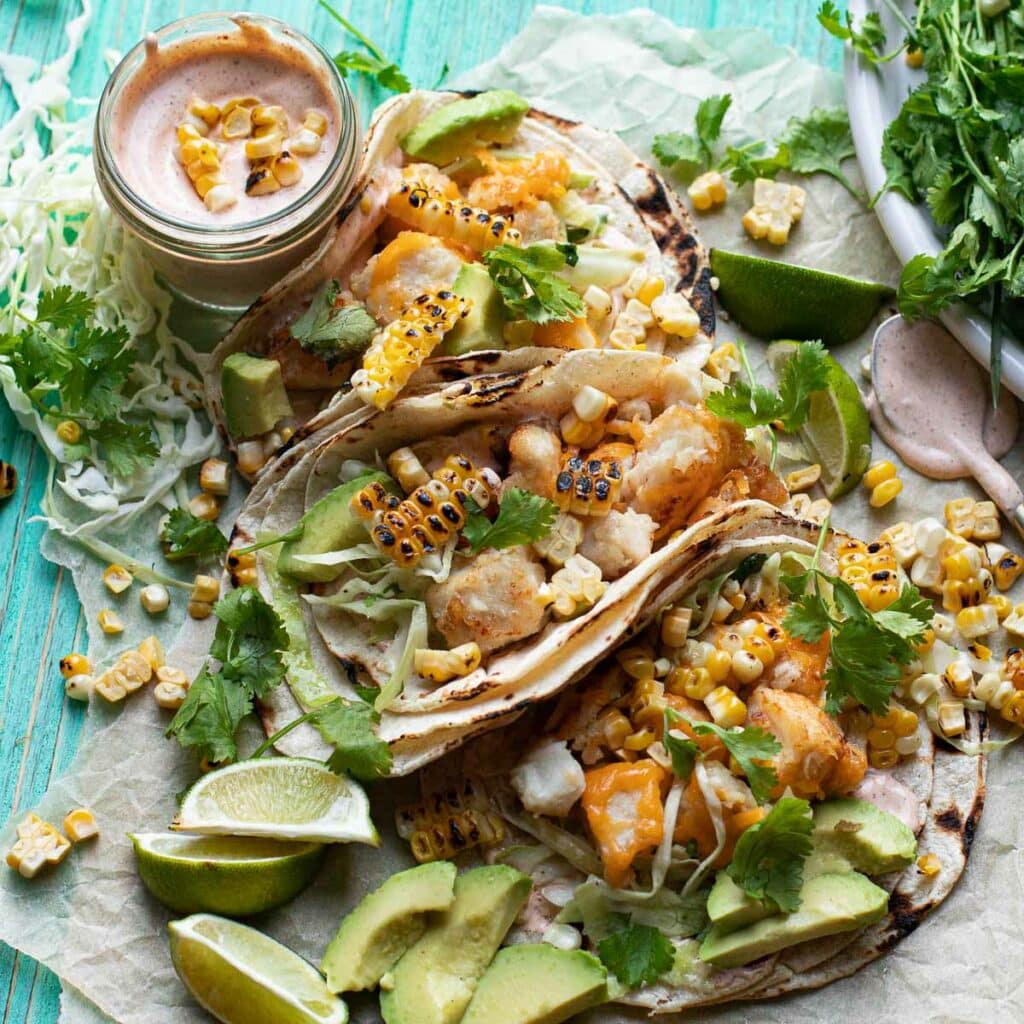 Three baja fish tacos garnished with corn, cilantro and limes
