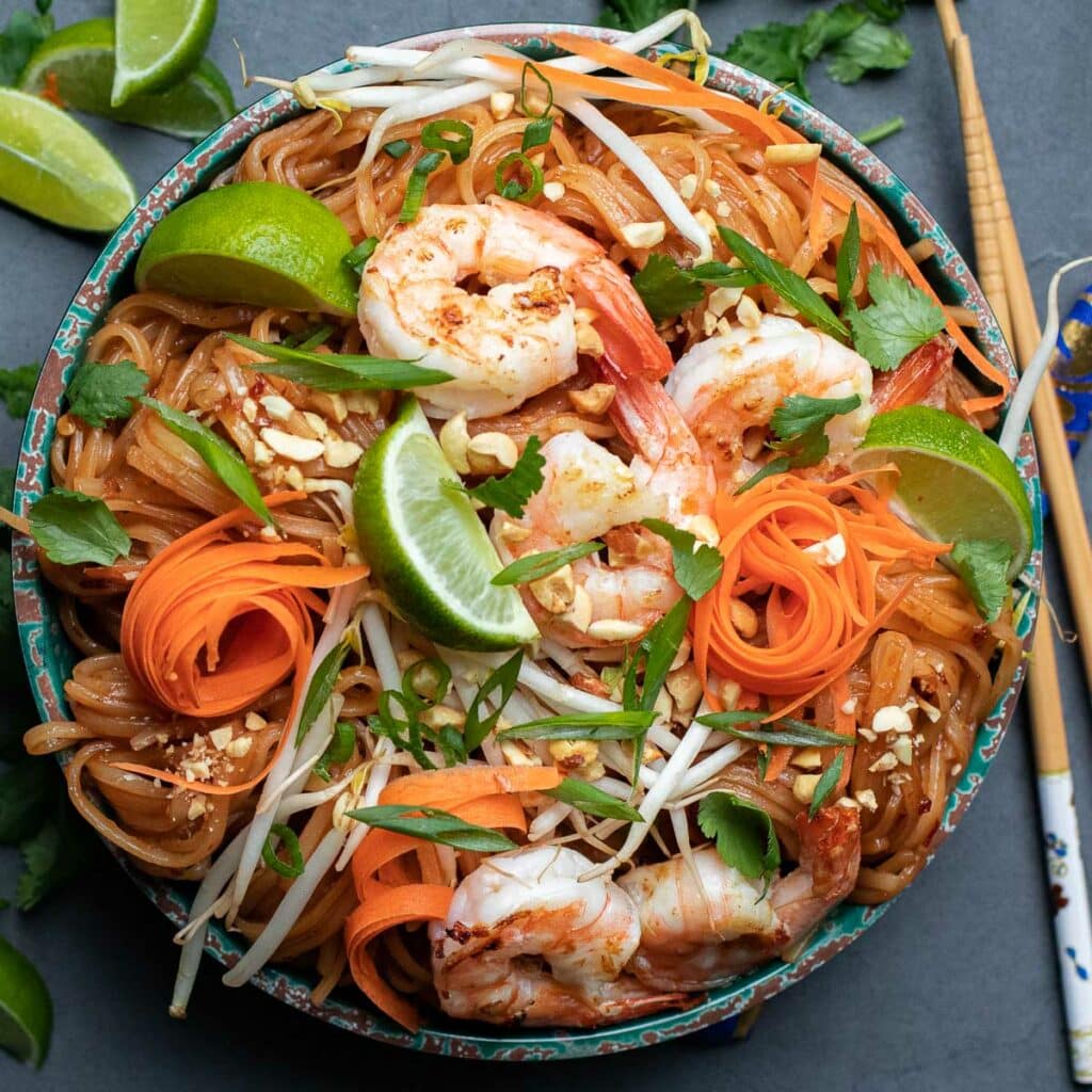 Bowl of pad thai noodles with shredded carrots, lime wedges, shrimp and bean sprouts