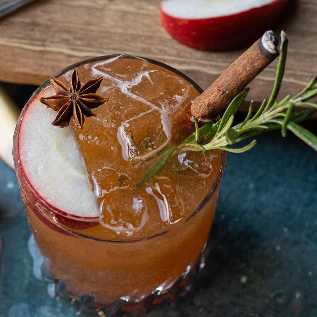 apple vodka cocktail in a rocks glass with an apple wedge, star anise, rosemary and cinnamon stick