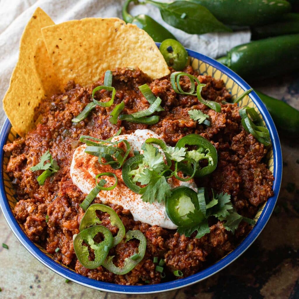 A bowl of Texas chili garnished with tortilla chips,  jalapeño peppers, scallions and cilantro