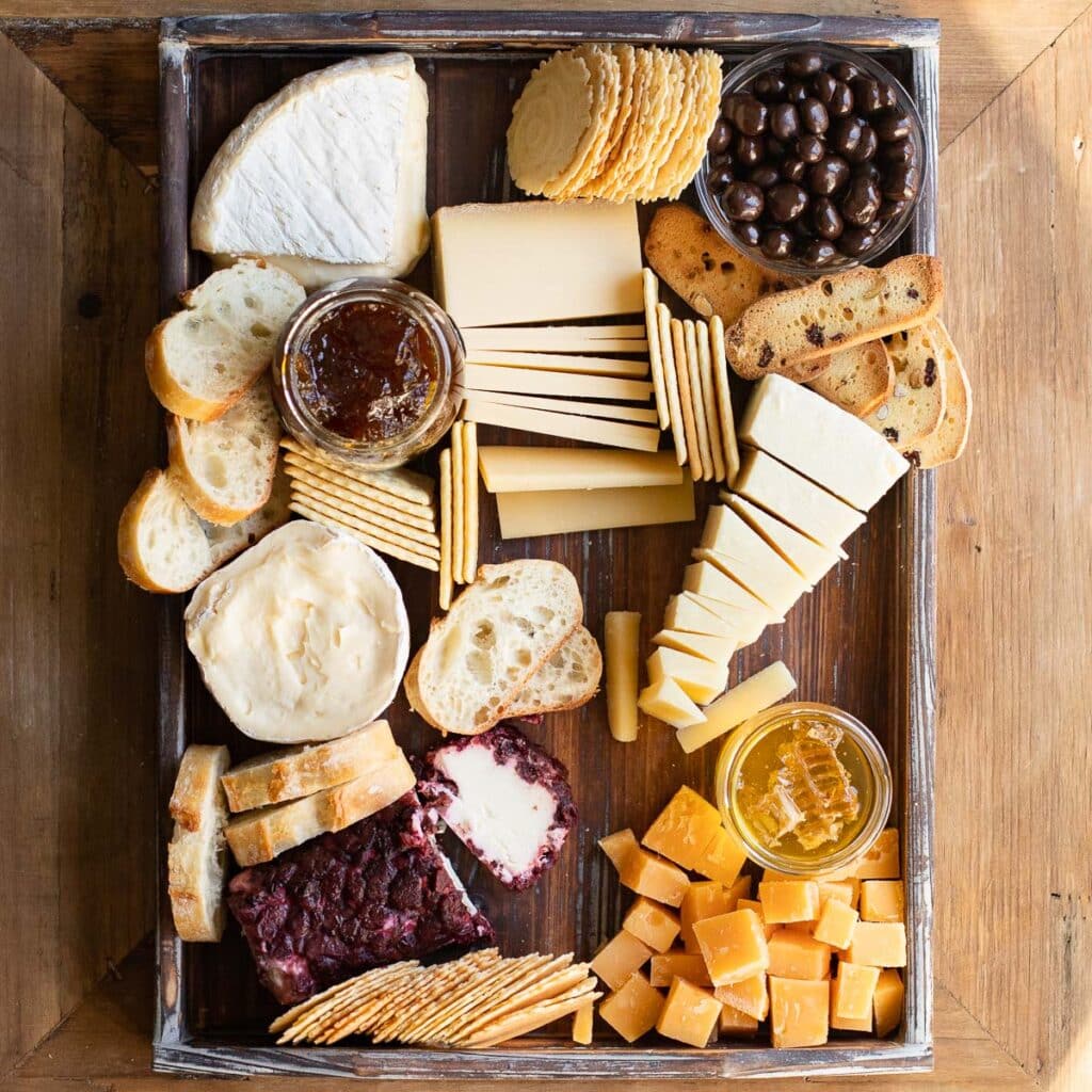 Cheeses. crackers, sliced baguette, a jar of honey and a jar of fig preserves on a wooden board