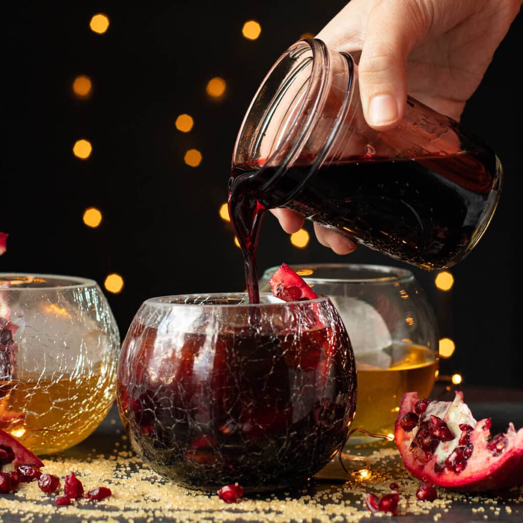 A hand pouring pomegranate syrup into a cocktail glass