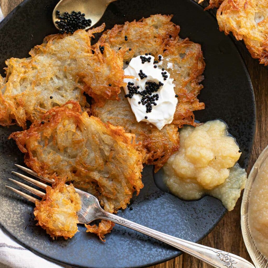 A pile of potato latkes on a black plate with sour cream, caviar and applesauce