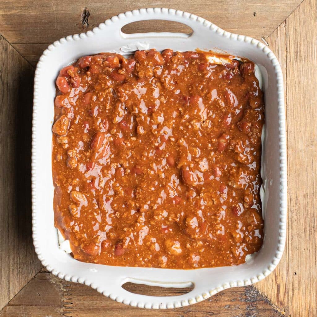 Canned chili spread across the cream cheese layer in a white baking dish