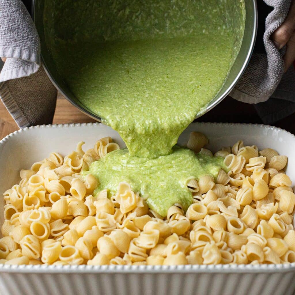 Pesto cream sauce being poured from a saucepan onto a baking dish full of cooked pasta
