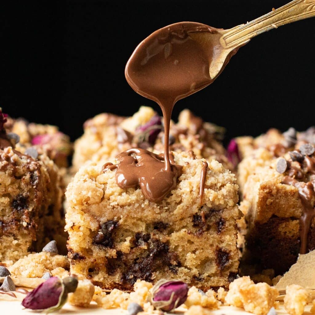 A gold spoon drizzling melted chocolate onto a piece of crumb cake