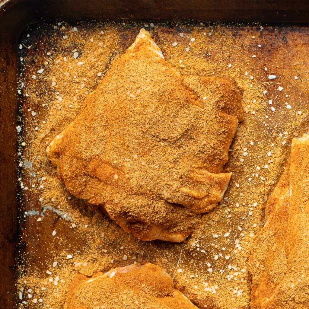 A piece of raw chicken thigh seasoned with a dry spice rub on baking sheet
