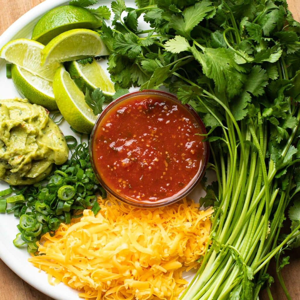 Fresh cilantro, lime wedges, avocado mash, salsa, chopped scallions and grated cheese arranged on a white plate