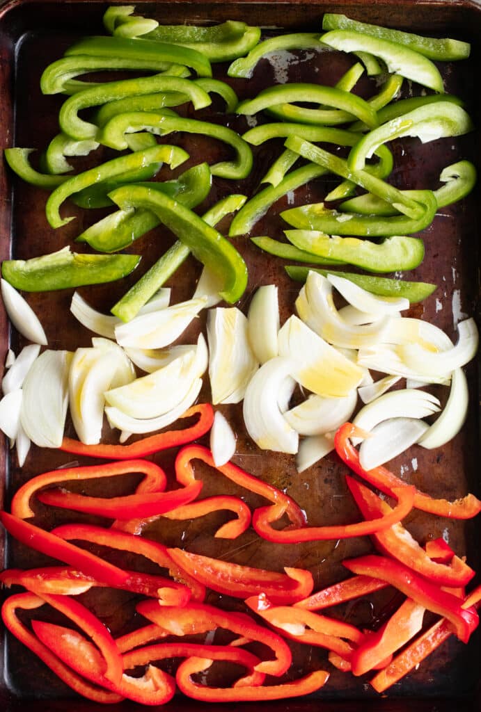 Sliced green bell peppers, sliced red bell peppers and sliced onions arranged on a baking sheet