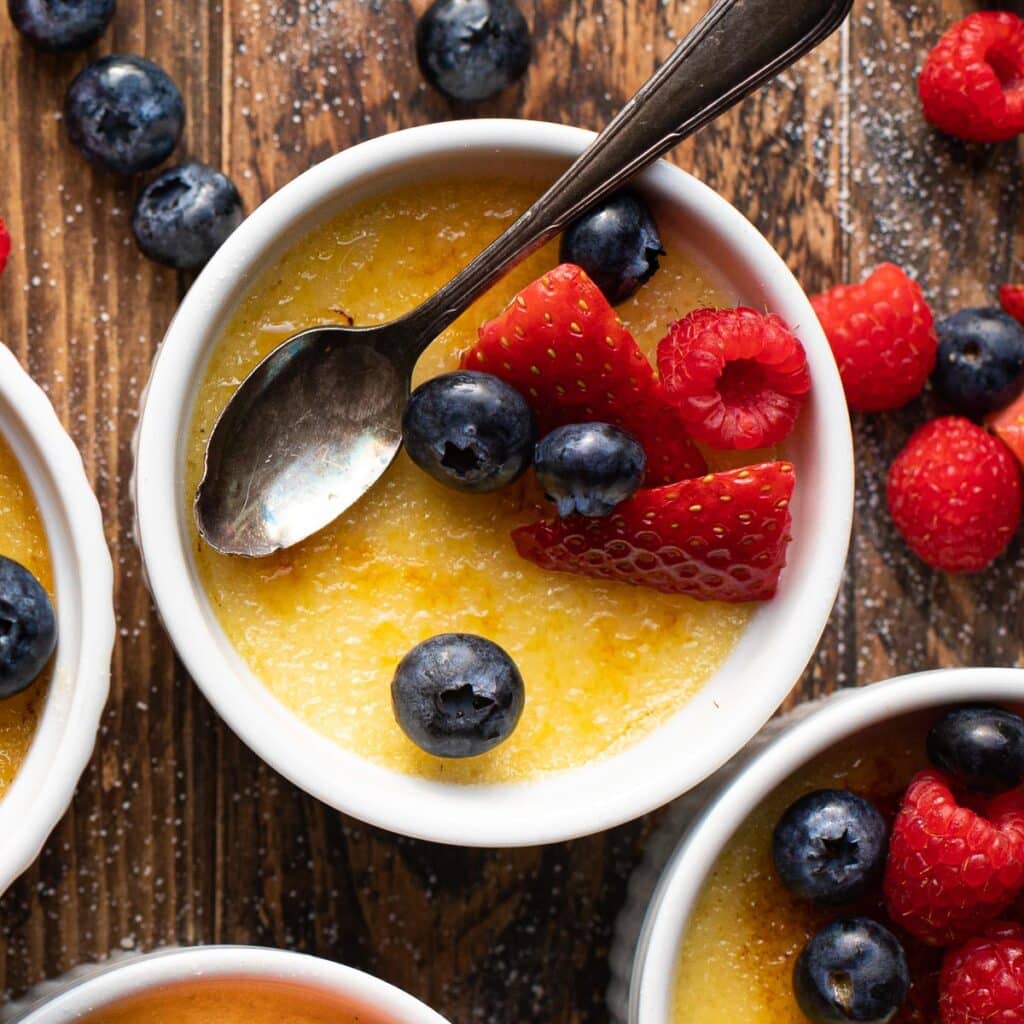 A white ramekin filled with creme brulee, topped with a silver spoon and berries.