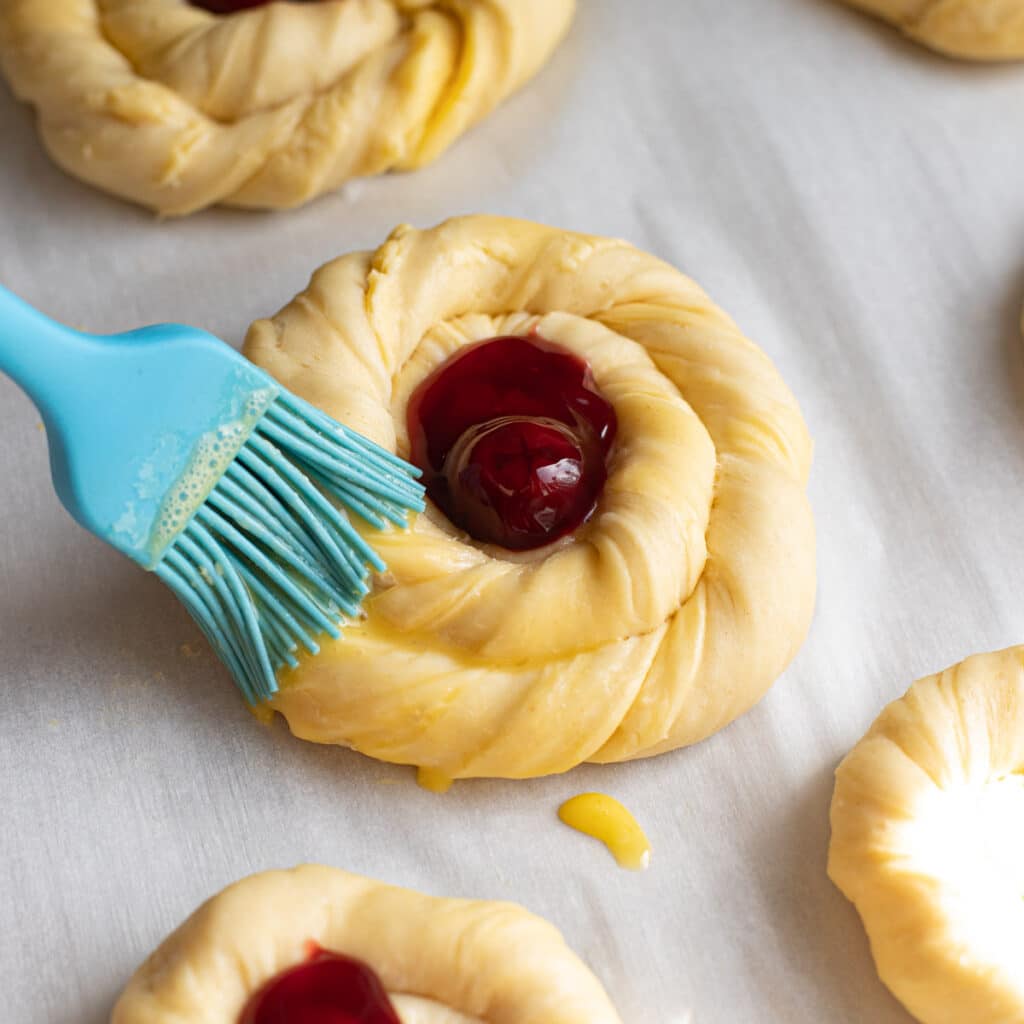 A cherry danish before baking being brushed with egg wash using a blue pastry brush