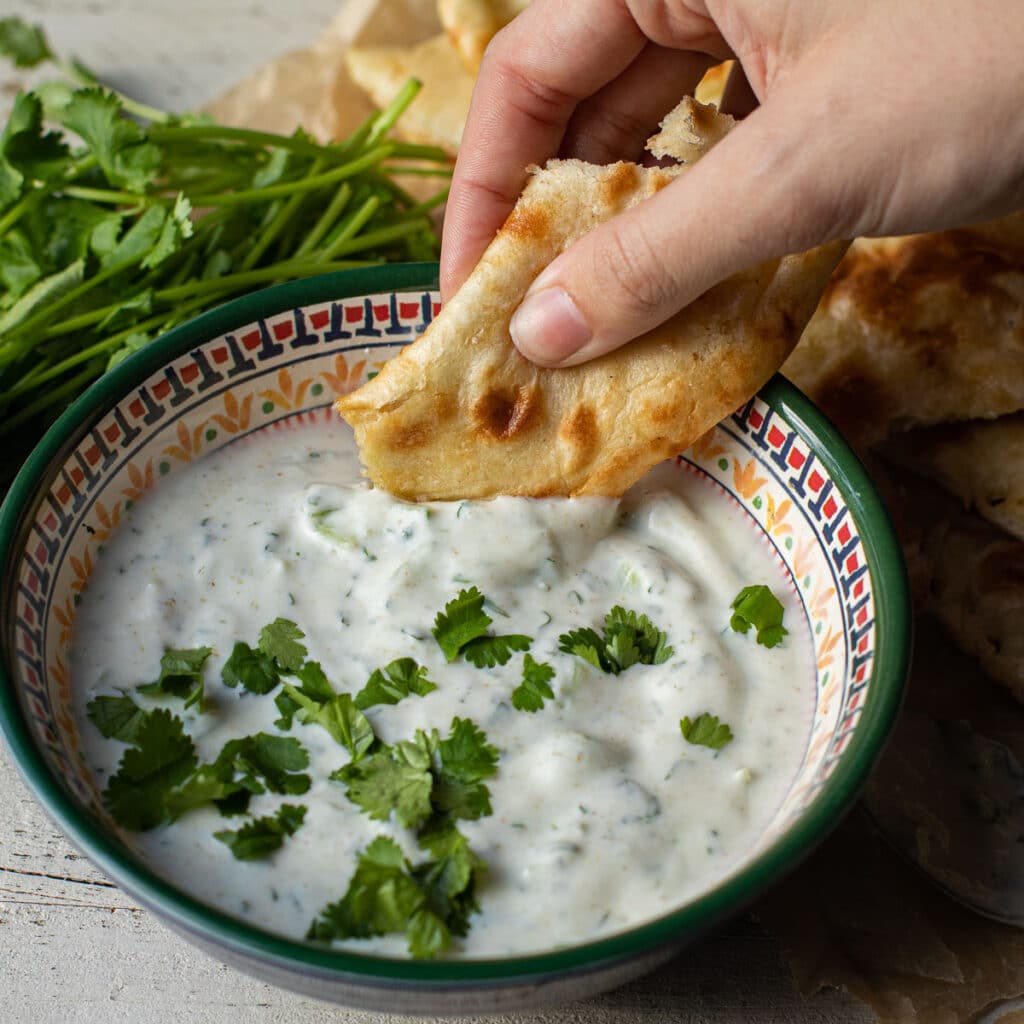 A piece of naan bread being dipped into a bowl of Indian yogurt sauce (raita)