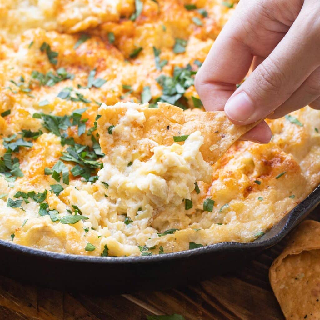 A hand dipping a chip into hot artichoke dip