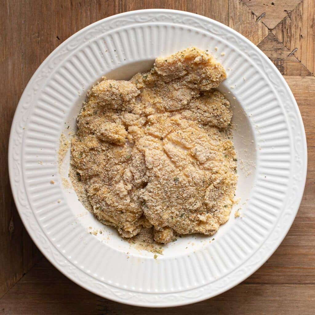 chicken breast being dredged in seasoned bread crumbs in a white bowl