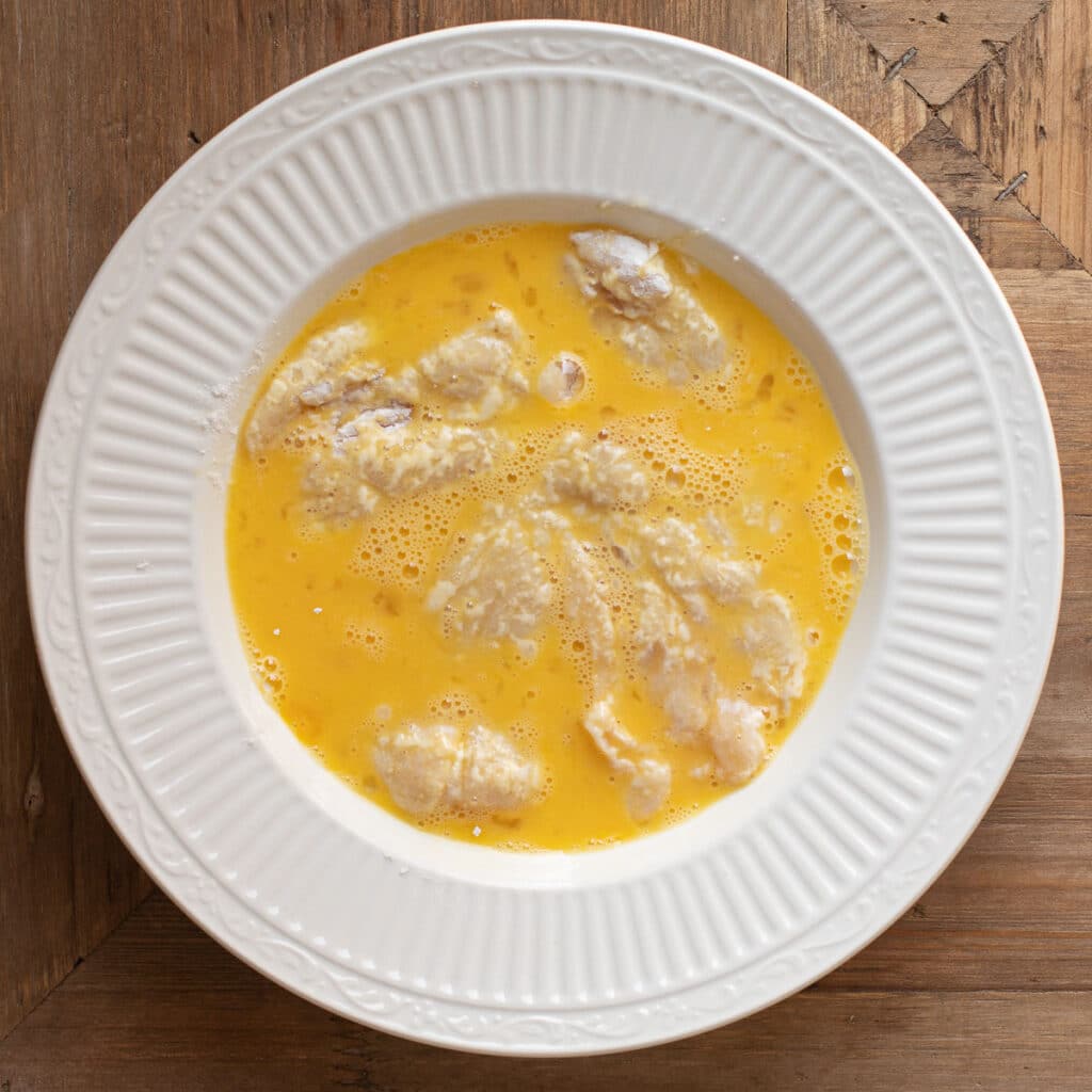 chicken breast being dredged in beaten egg mixture in a white bowl