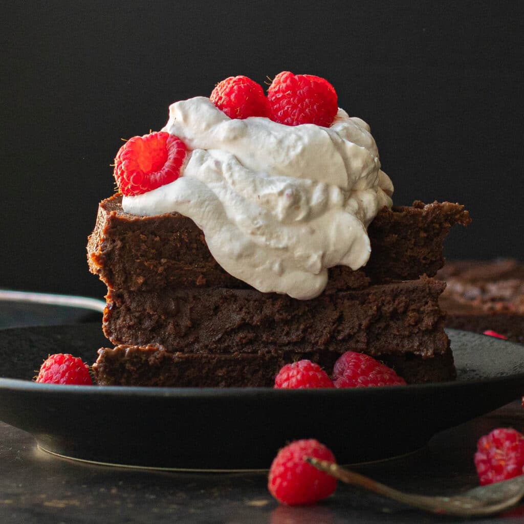 3 slices of flourless chocolate cake stacked up and topped with whipped cream and raspberries