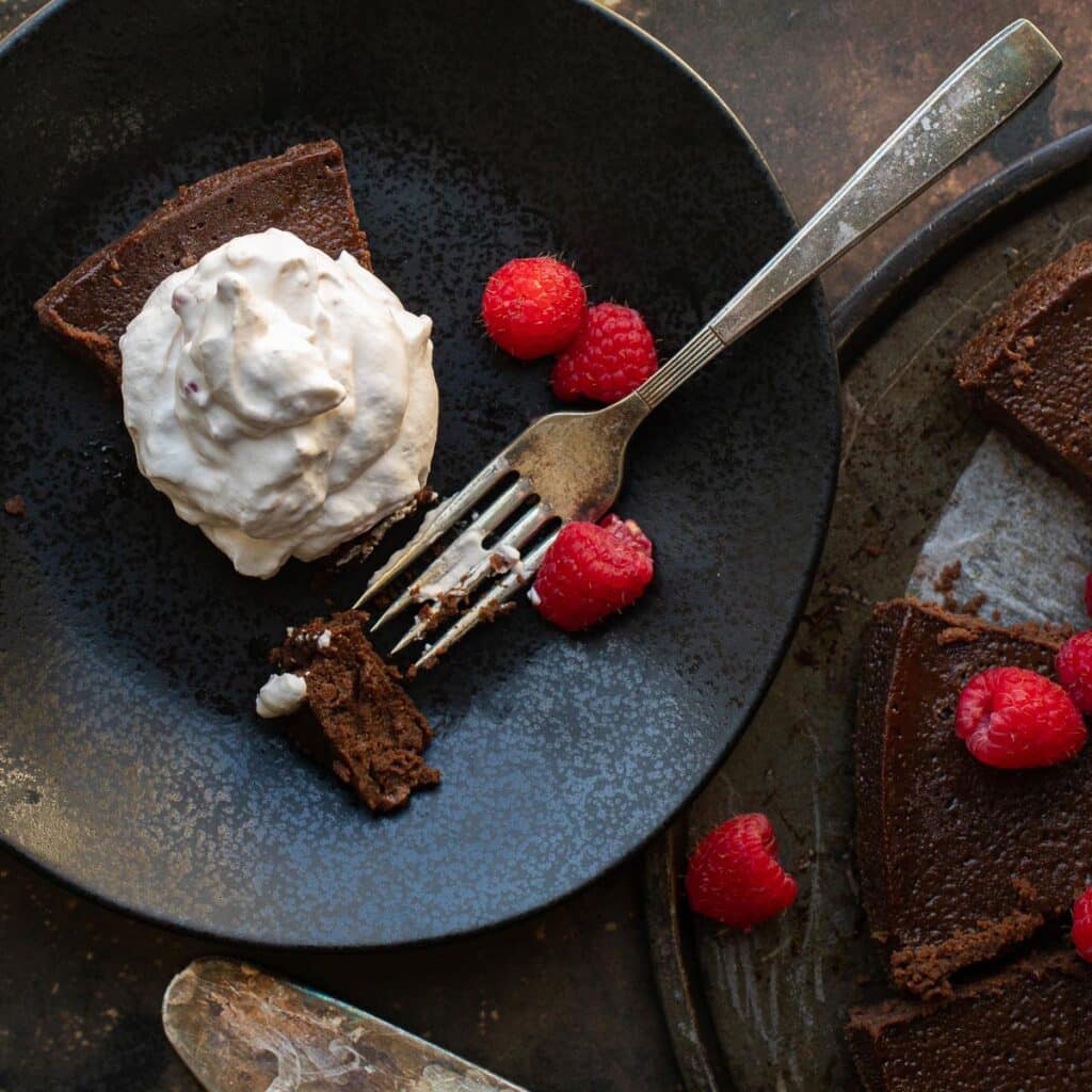 a slice of flourless chocolate cake on a black plate, topped with whipped cream and raspberries