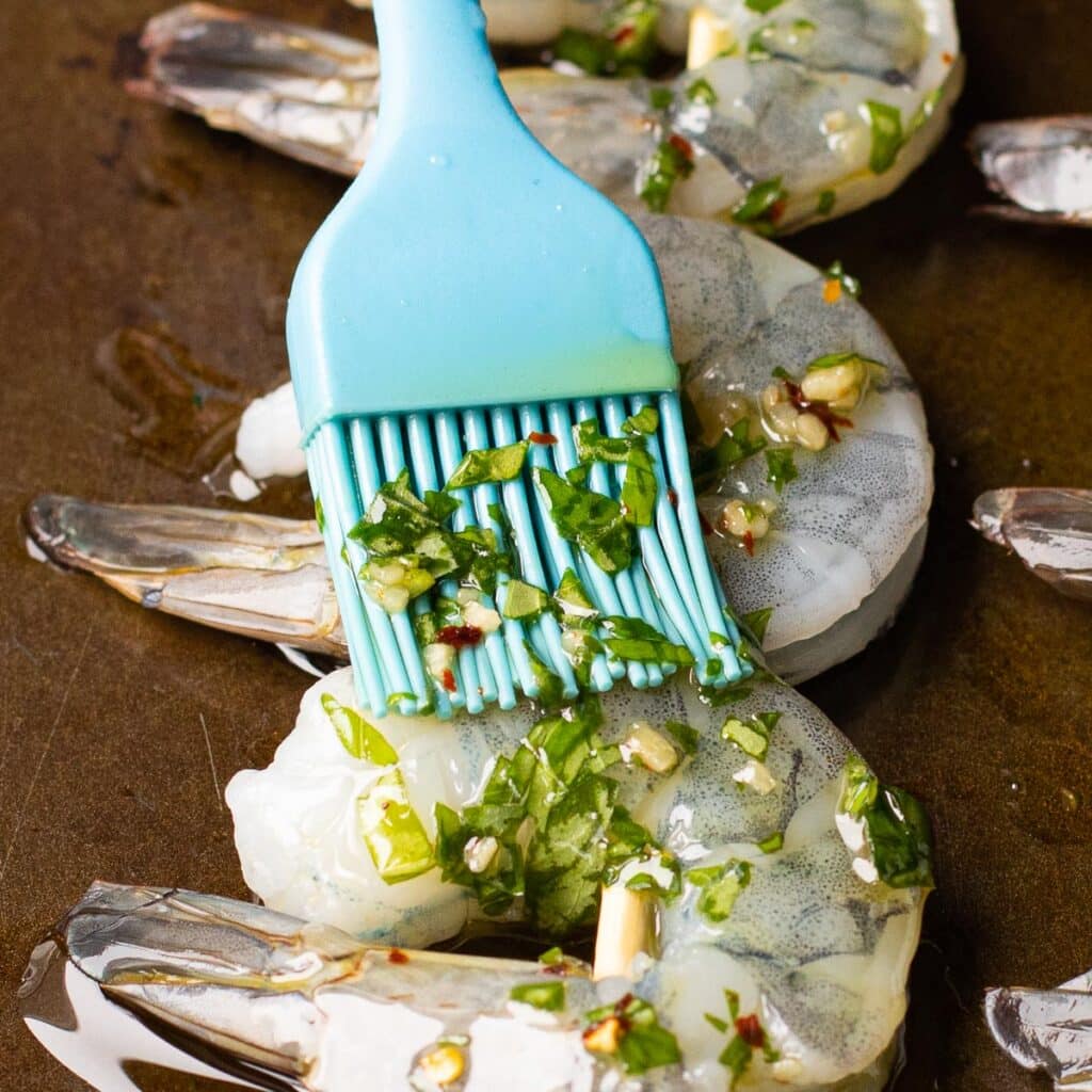 A blue silicone brush brushing herb oil onto raw shrimp
