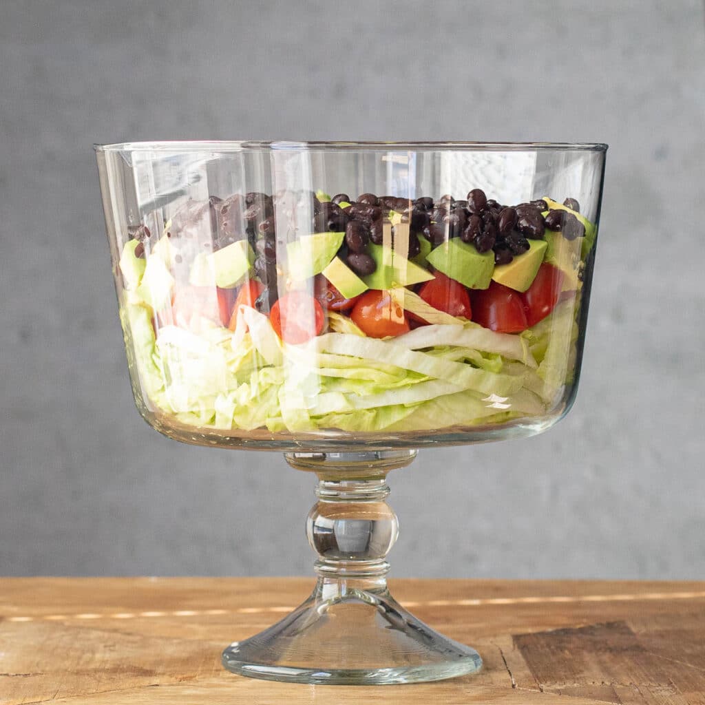 layers of lettuce, tomato, avocado and black beans in a glass trifle bowl