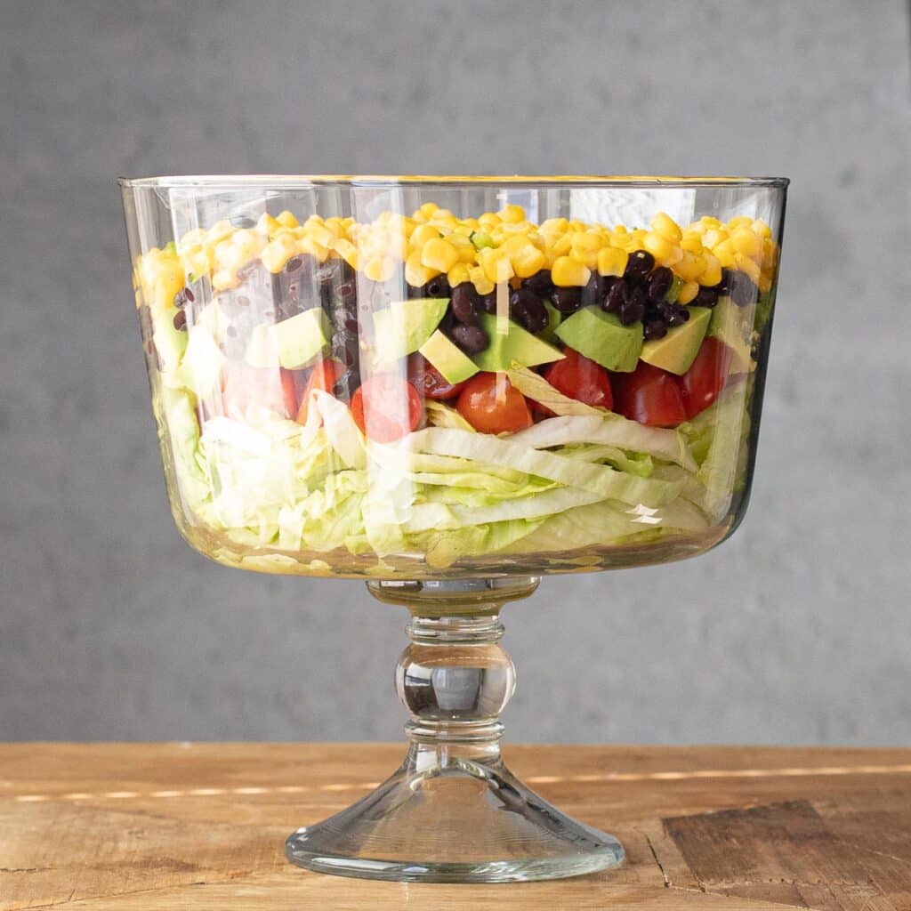 layers of lettuce, tomato, avocado, black beans and corn in a glass salad bowl
