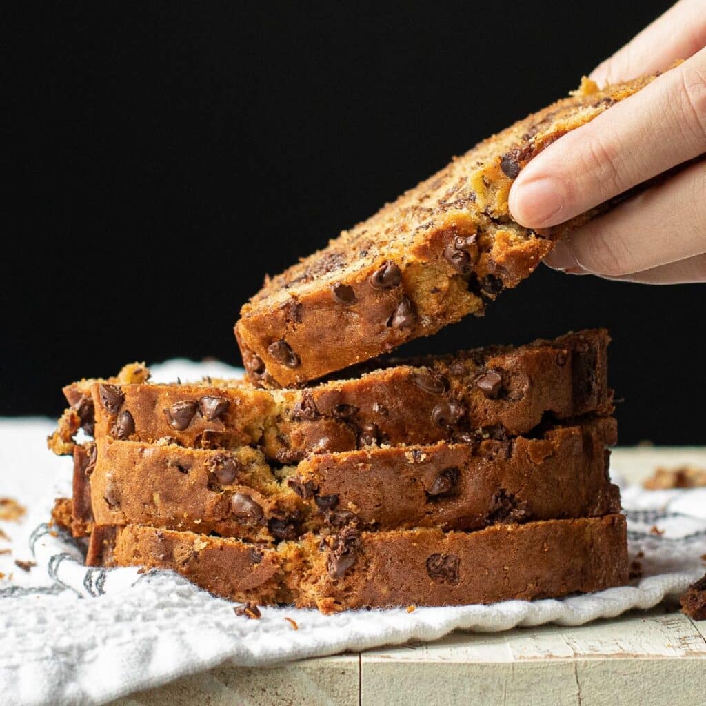 a hand lifting a slice of chocolate chip banana bread from a stack of slices