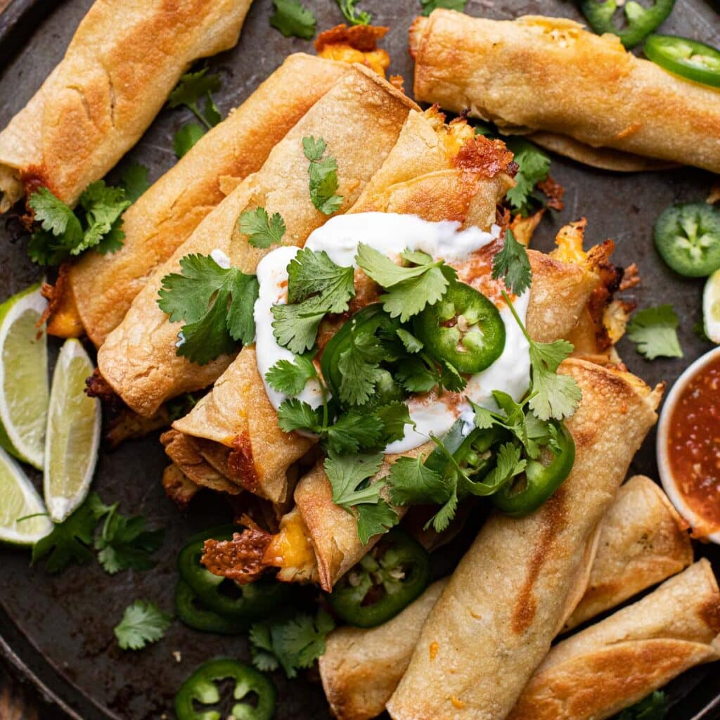 a stack of baked taquitos topped with sour cream, cilantro and peppers