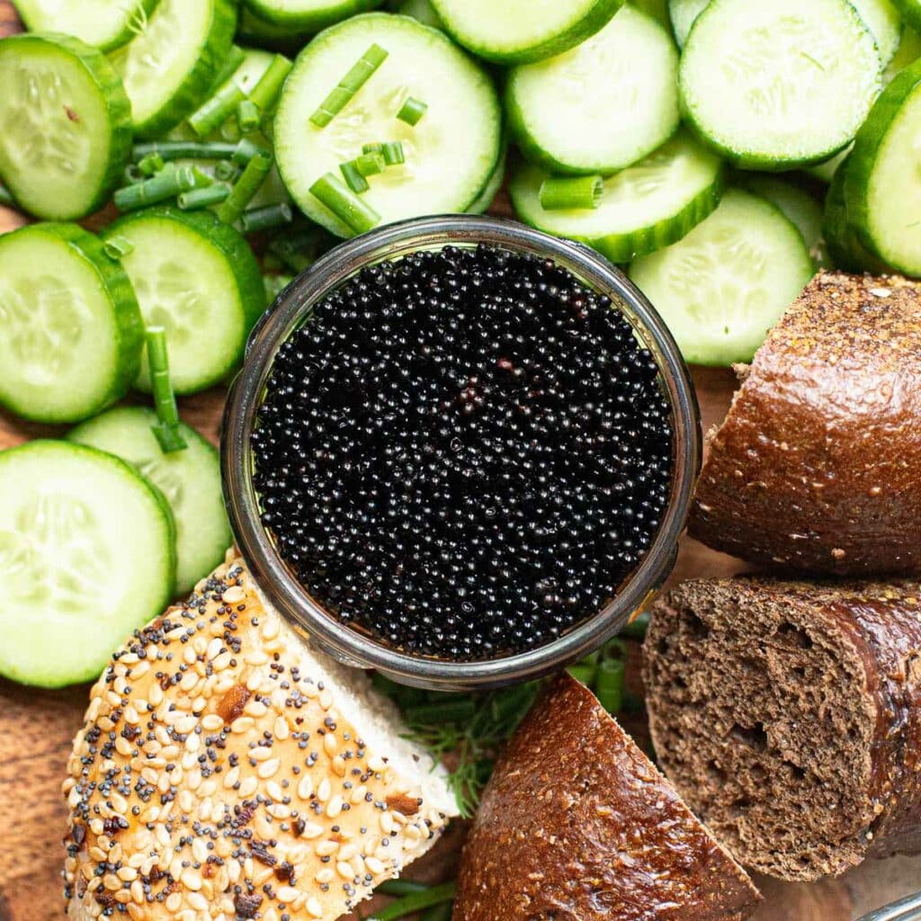 black caviar in a glass bowl surrounded by bagels and sliced cucumber