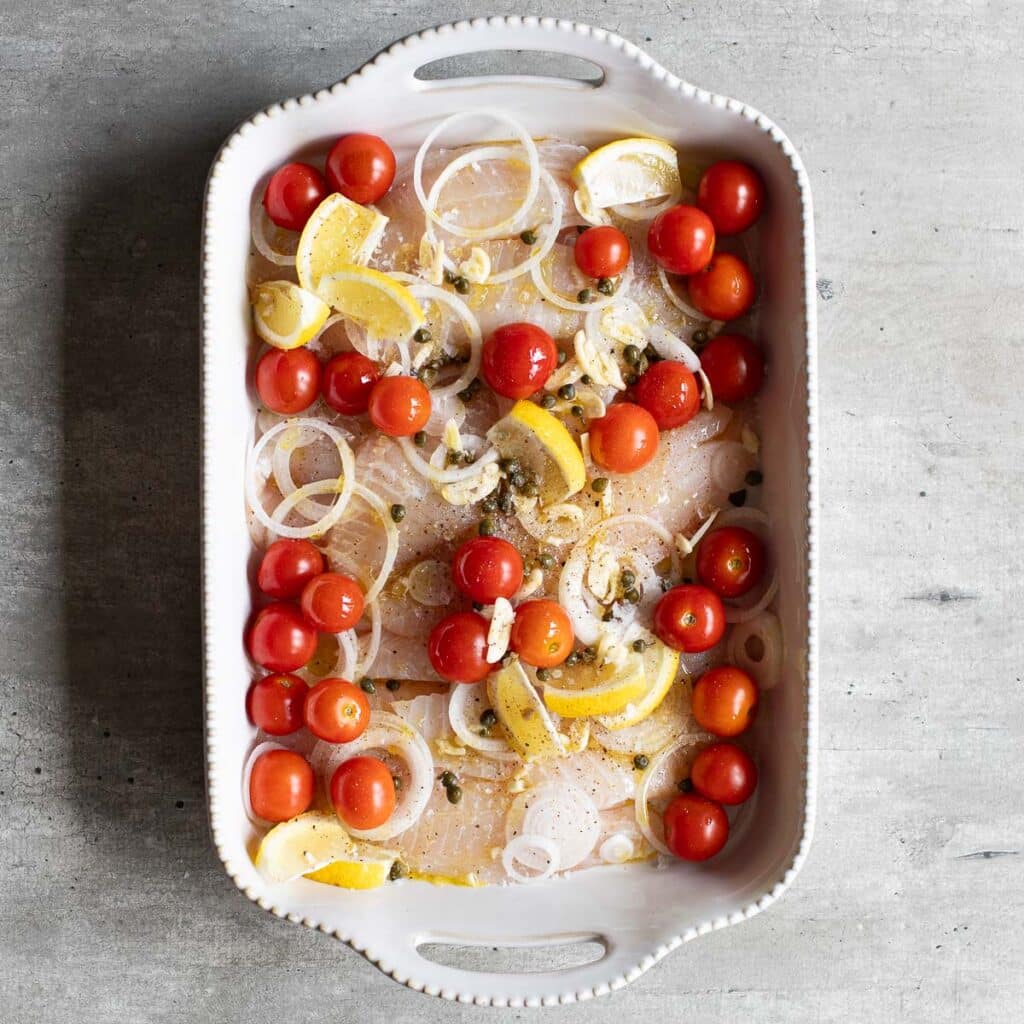white tilapia fish in a large baking dish with lemon wedges, sliced onions and cherry tomatoes before it is roasted