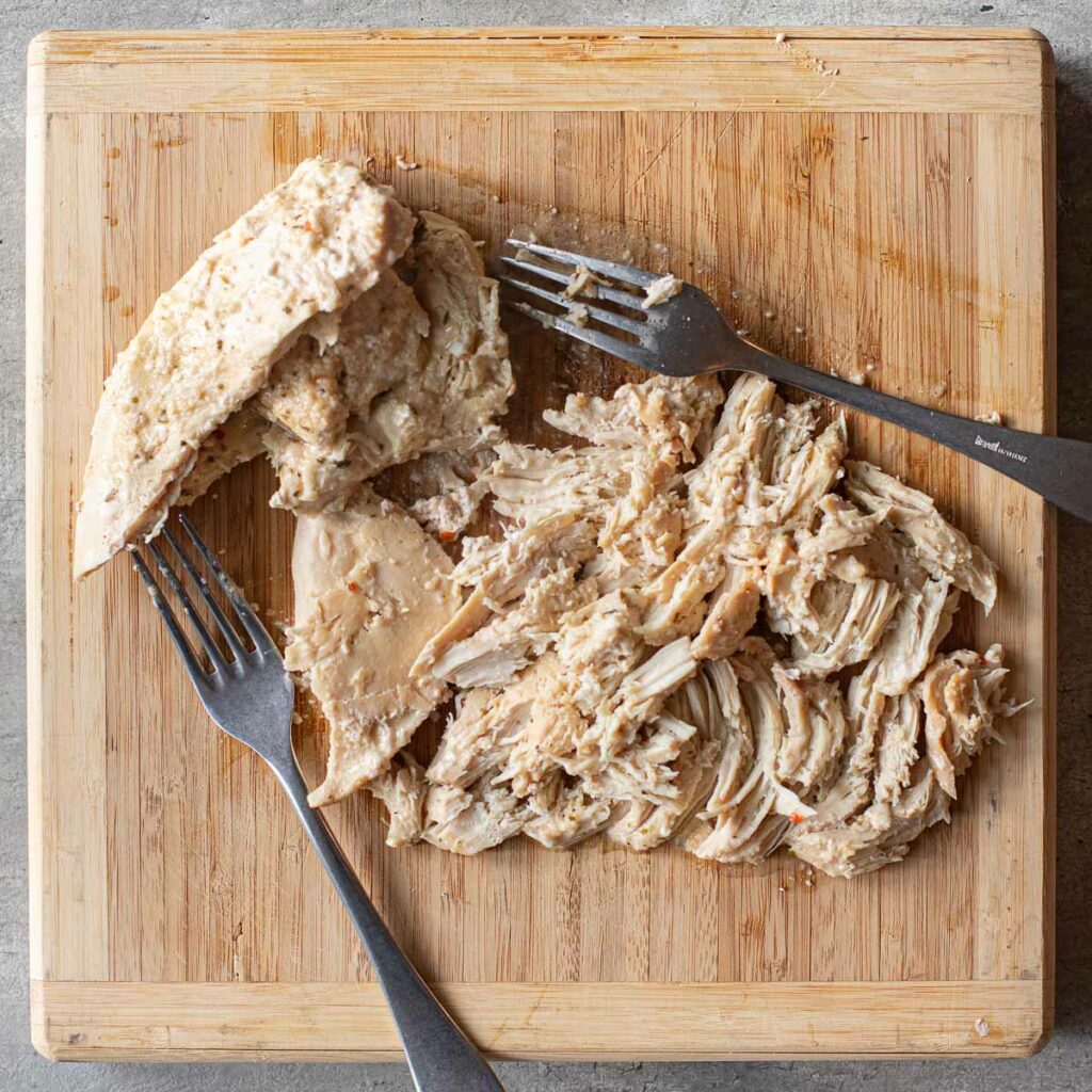 cooked chicken breasts being shredded with 2 forks on a wooden board