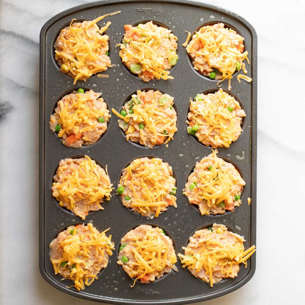 uncooked chicken muffins topped with grated cheese in a muffin pan