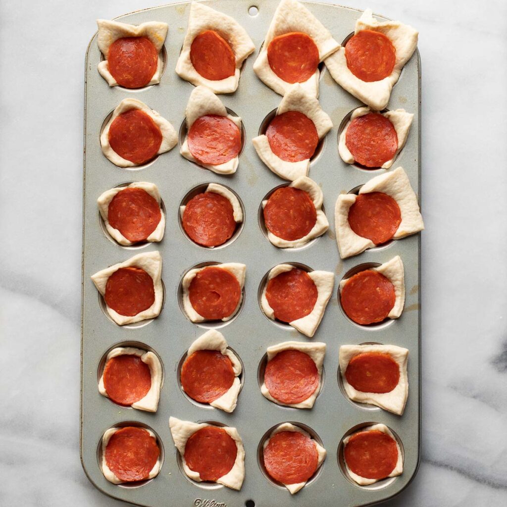 slices of pepperoni and squares of crescent roll dough in a mini muffin pan