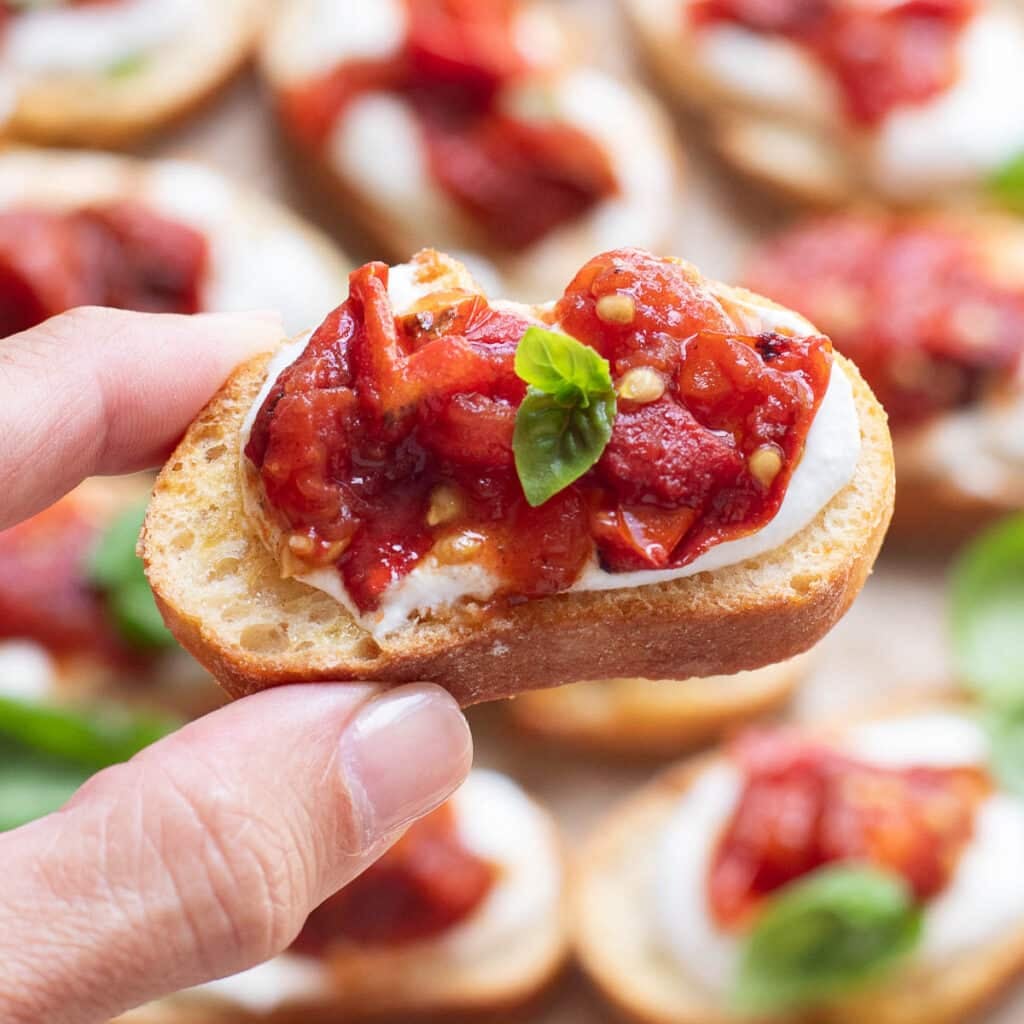 a hand holding a piece of cottage cheese toast topped with roasted tomatoes and fresh basil