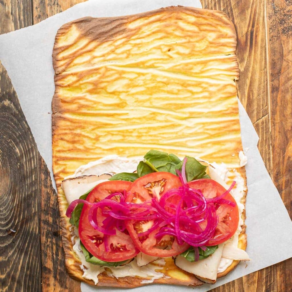 A cottage cheese wrap, laid flat on a wooden board with half covered in deli turkey, lettuce, tomato and pickled onions.