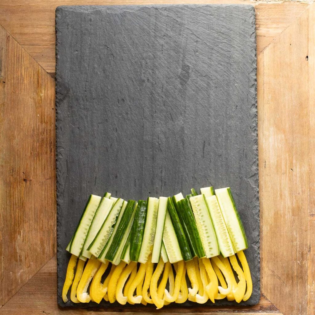 cucumber strips overlapping yellow bell pepper strips on a black slate board
