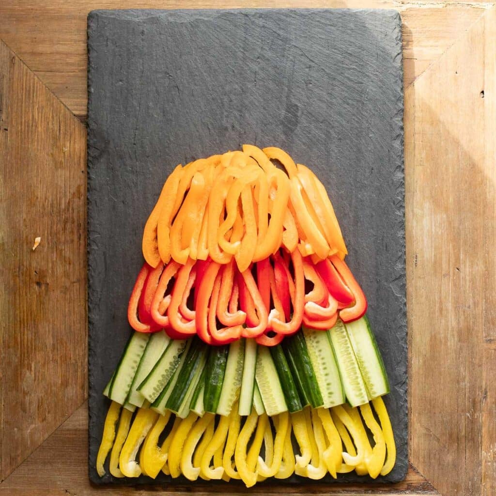 orange bell pepper strips, red bell pepper strips, cucumber strips and yellow bell pepper strips arranged in an overlapping pattern on a black slate board