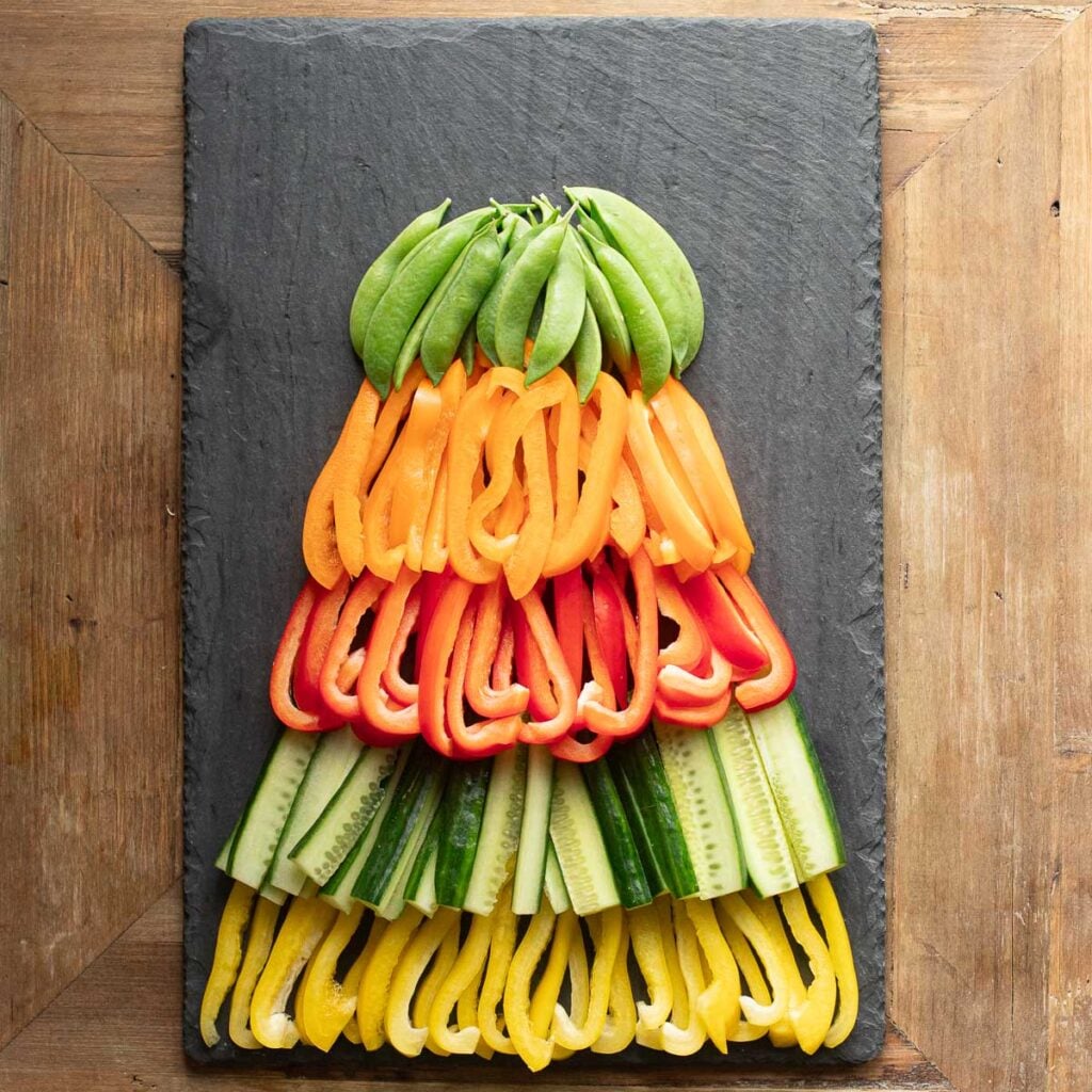 snow peas, orange bell peppers, red bell peppers, cucumber strips and yellow bell peppers arranged in a tree shape on a black slate board
