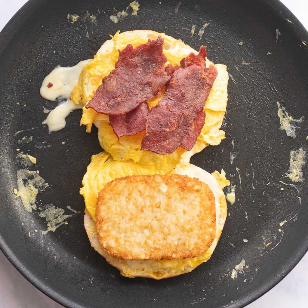 strips of bacon, a hashbrown patty and eggs on halves of a bagel in a pan