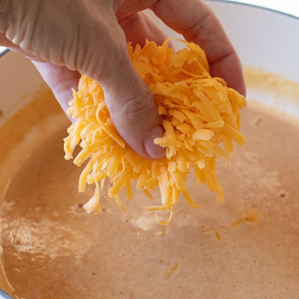 a hand putting grated cheese into a pot of melted cheese
