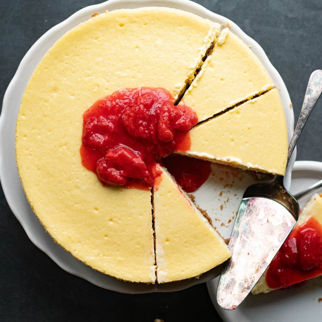 overhead view of a whole New York Cheesecake with a few slices made, topped with strawberry compote