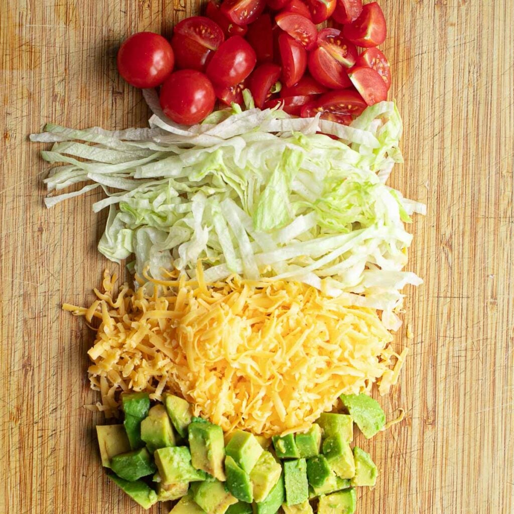 chopped tomatoes, shredded lettuce, grated cheddar cheese and diced avocado on a wooden cutting board