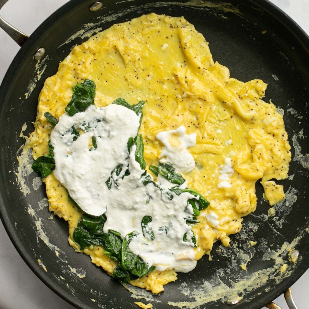 omelet in a pan with half of the eggs covered with cottage cheese and spinach
