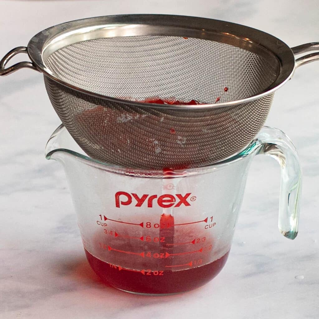 strawberry syrup being strained into a glass measuring cup